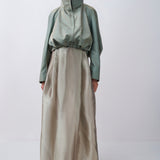 A model stands against a light gray studio background wearing a long-sleeved, high-neck, floor-length dress with a sage green upper section and a flowing champagne-beige skirt.