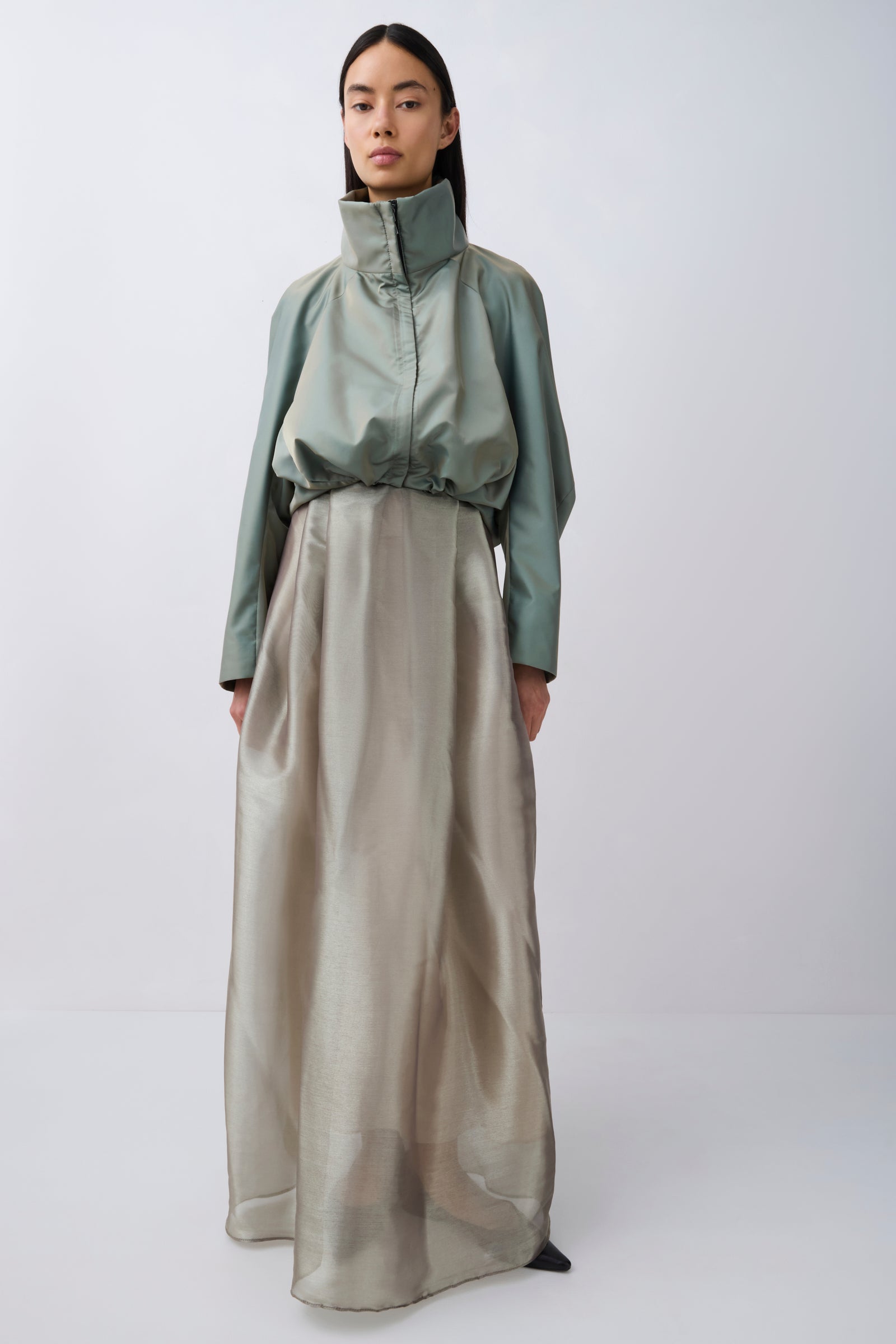 A model stands against a light gray studio background wearing a long-sleeved, high-neck, floor-length dress with a sage green upper section and a flowing champagne-beige skirt.