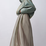 A model wearing a futuristic, voluminous two-piece outfit featuring a high-collared sage green jacket and a matching long, flowing skirt in a light taupe-beige color, set against a plain light gray studio background.