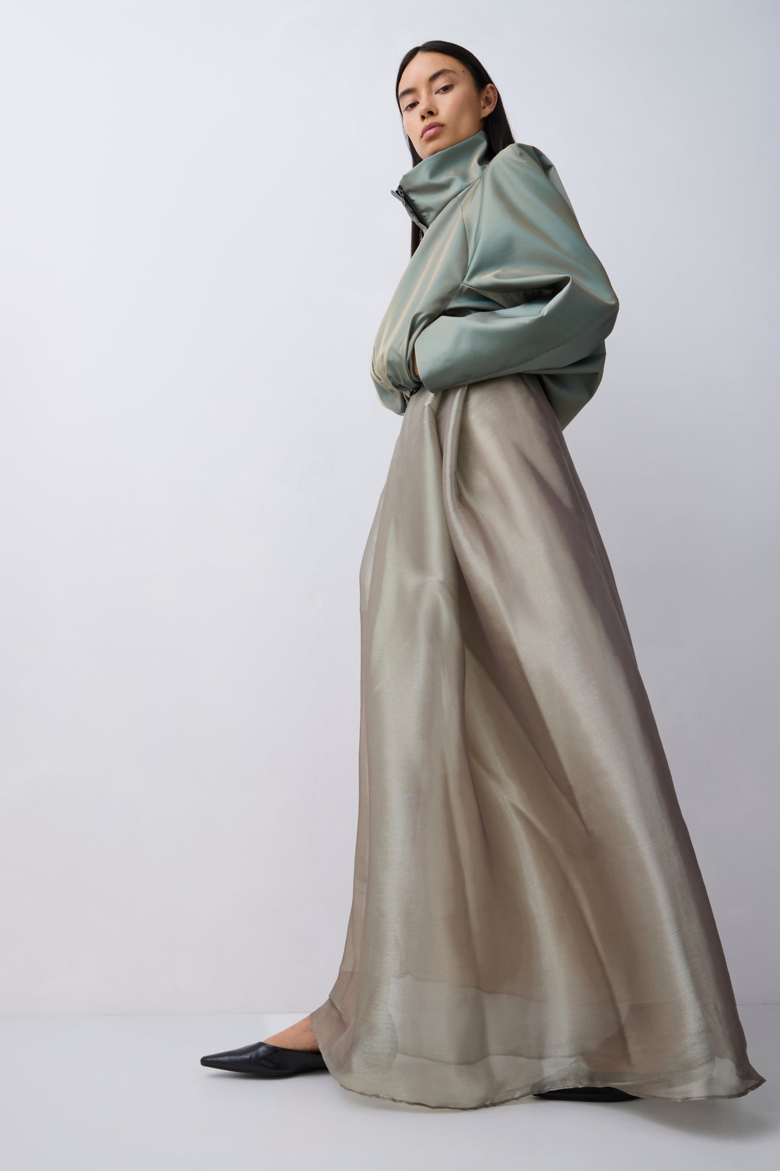 A model wearing a high-neck, voluminous sage green jacket and a long, flowing taupe-beige skirt, styled with black pointed flats against a light gray studio background.