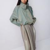 A model wearing a sage green high-neck long-sleeve jacket paired with a metallic taupe satin midi skirt, standing against a plain light gray studio background.
