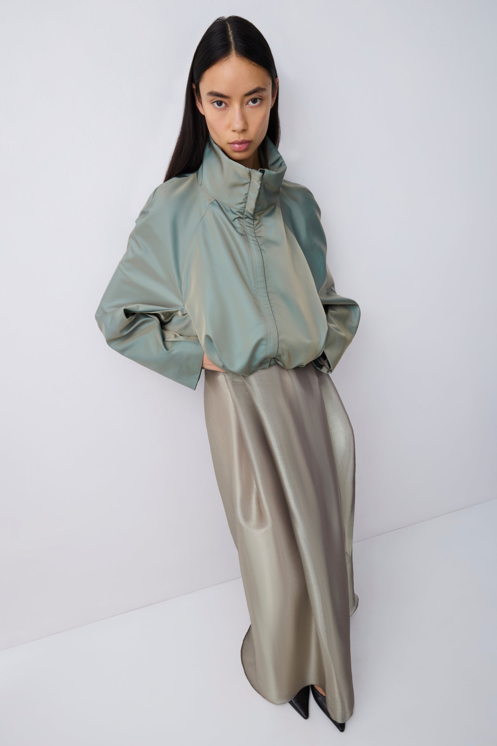 A model wearing a sage green high-neck long-sleeve jacket paired with a metallic taupe satin midi skirt, standing against a plain light gray studio background.