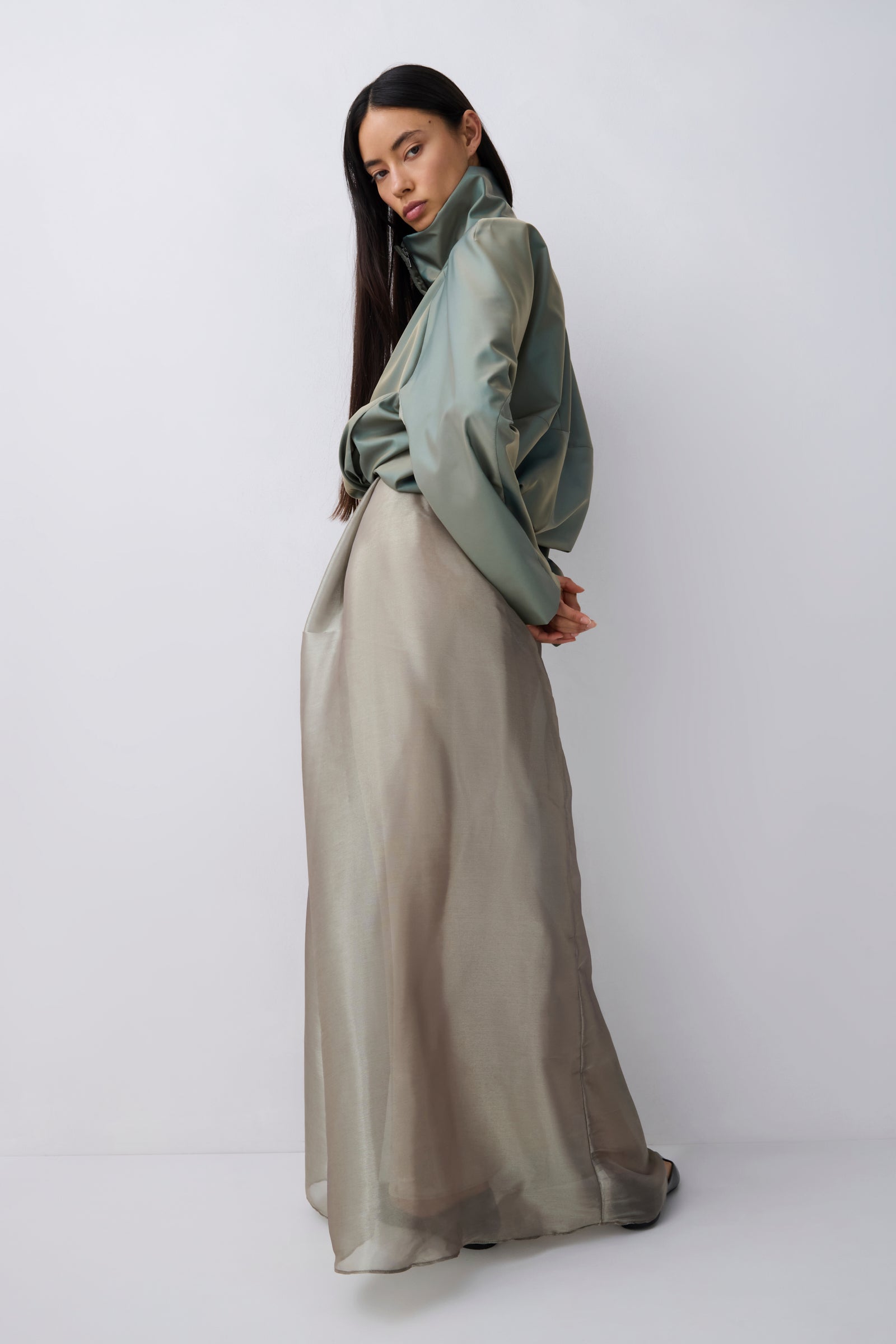 A model wearing a sage green high-neck long-sleeve blouse and a matching taupe-beige maxi skirt, standing against a light gray studio background.