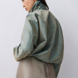 A model shown from a side-back angle wearing a voluminous sage green jacket and a matching taupe skirt against a plain light gray studio background.
