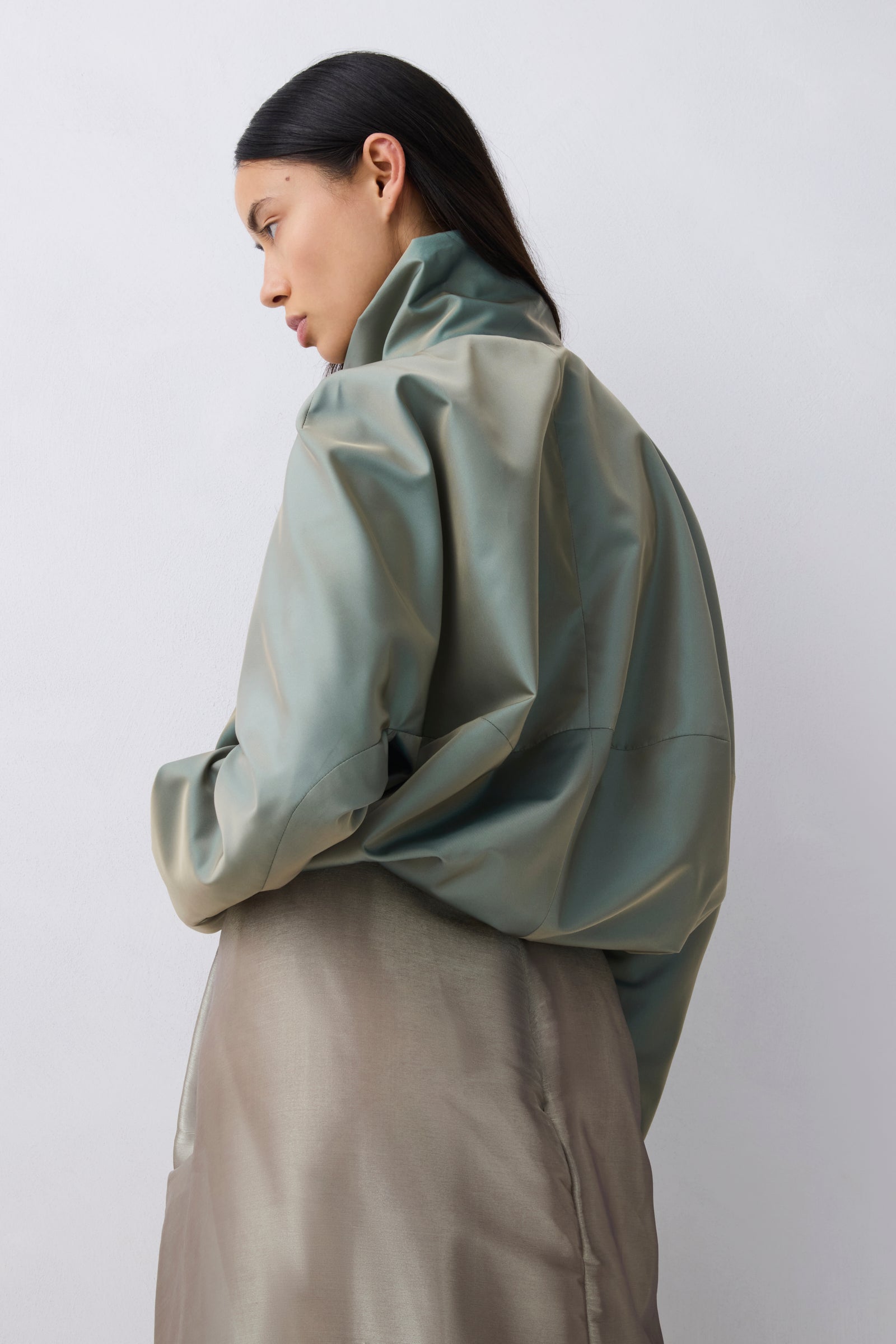 A model shown from a side-back angle wearing a voluminous sage green jacket and a matching taupe skirt against a plain light gray studio background.