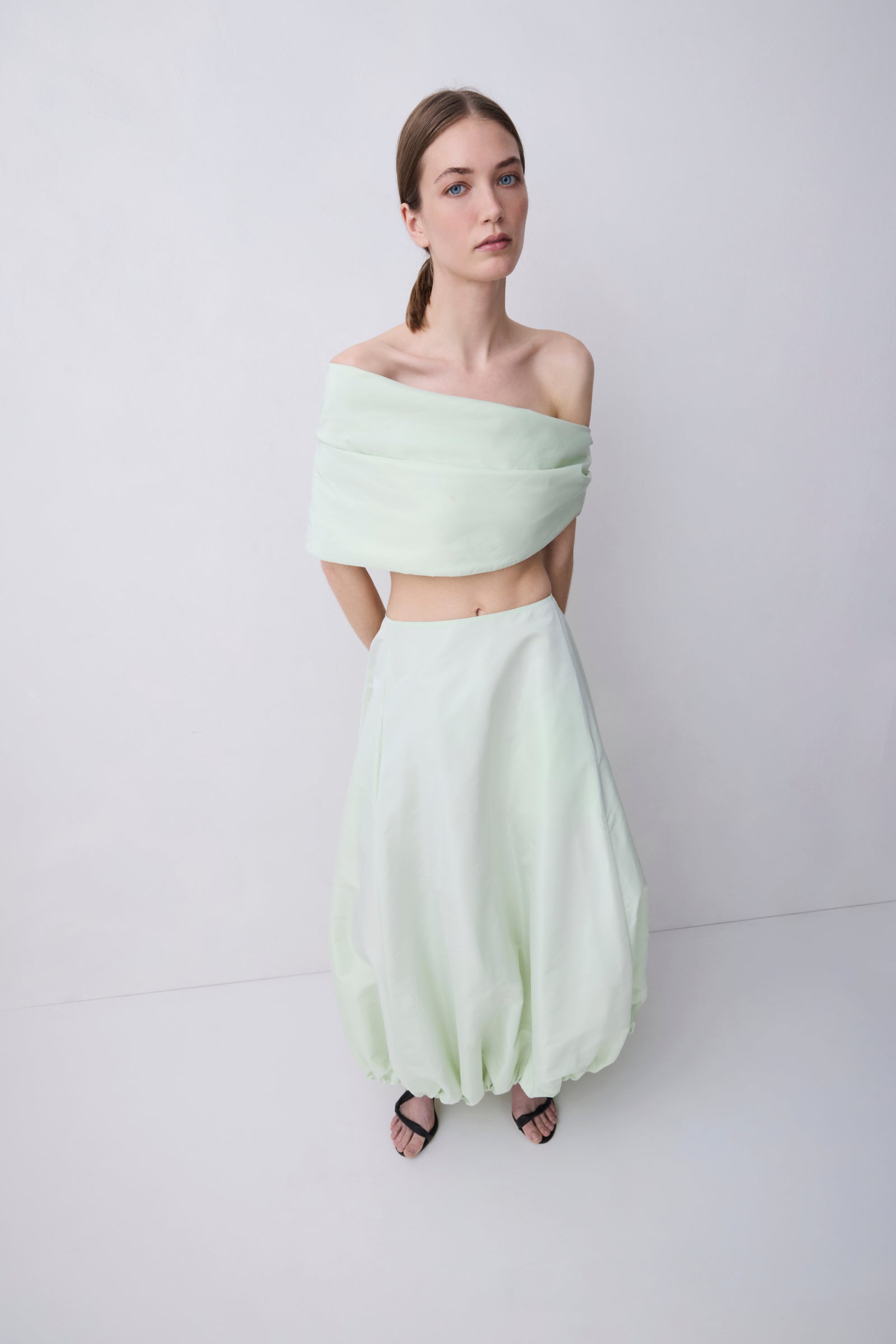 A model wearing a pale mint green two-piece outfit consisting of an off-the-shoulder wrap-style crop top and matching voluminous balloon skirt, paired with black sandals, against a plain light gray studio background.