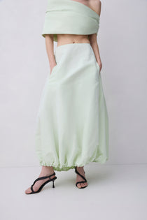 A model wearing a light sage green off-the-shoulder crop top and matching high-waisted, voluminous maxi skirt with black strappy high-heeled sandals, set against a plain light gray studio background.