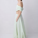 A woman stands in profile wearing a pale mint green two-piece outfit with an off-the-shoulder wrap-style crop top and a matching long skirt, paired with black strappy heels, against a plain light gray studio background.