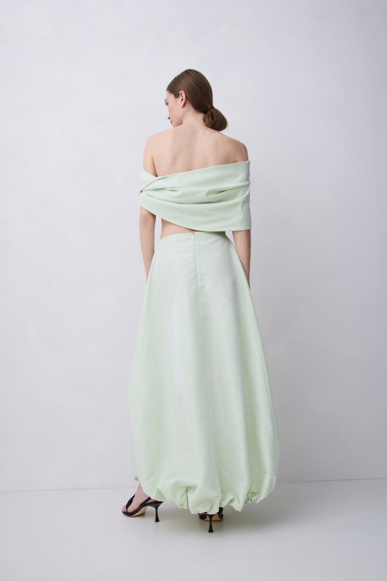 A woman stands with her back to the camera wearing a pale mint green off-the-shoulder maxi dress with a voluminous gathered skirt and black high-heeled sandals, set against a plain light gray studio background.