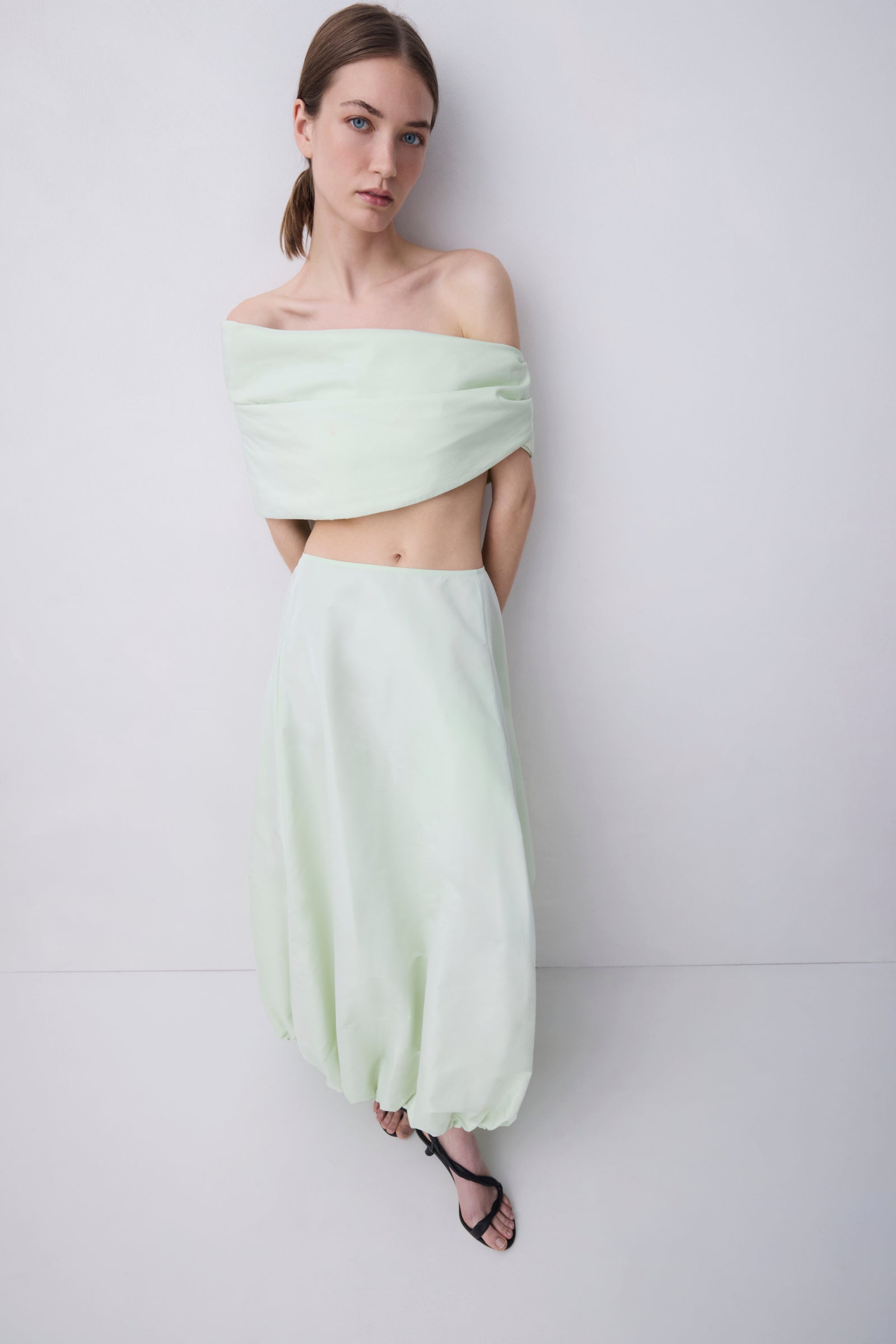 A model wearing a light mint green two-piece set consisting of an off-the-shoulder crop top and a matching high-waisted, voluminous maxi skirt, paired with black strappy sandals against a plain light gray studio background.