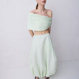 A model wearing a pale mint green two-piece outfit consisting of an off-the-shoulder wrap-style crop top and matching voluminous balloon skirt, paired with black sandals, against a plain light gray studio background.