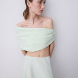 A woman modeling a pale mint green two-piece outfit consisting of an off-the-shoulder wrap-style crop top and a matching high-waisted skirt, set against a plain light gray studio background.
