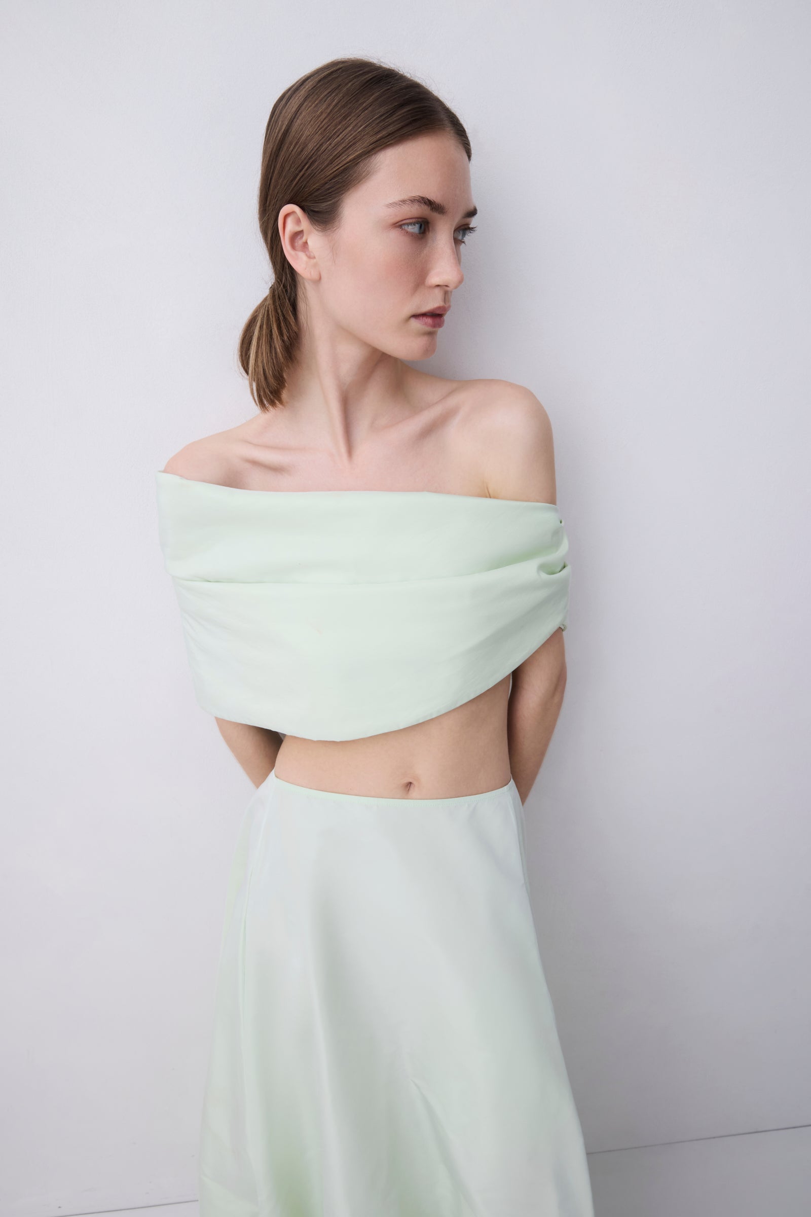 A woman modeling a pale mint green two-piece outfit consisting of an off-the-shoulder wrap-style crop top and a matching high-waisted skirt, set against a plain light gray studio background.
