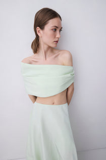A woman modeling a pale mint green two-piece outfit consisting of an off-the-shoulder wrap-style crop top and a matching high-waisted skirt, set against a plain light gray studio background.