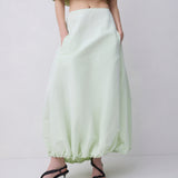 A model wearing a light sage green off-the-shoulder crop top and matching high-waisted, voluminous maxi skirt with black strappy high-heeled sandals, set against a plain light gray studio background.