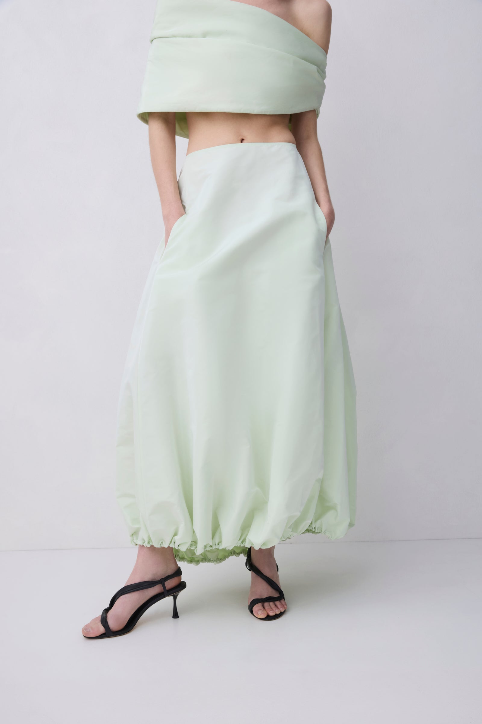 A model wearing a light sage green off-the-shoulder crop top and matching high-waisted, voluminous maxi skirt with black strappy high-heeled sandals, set against a plain light gray studio background.