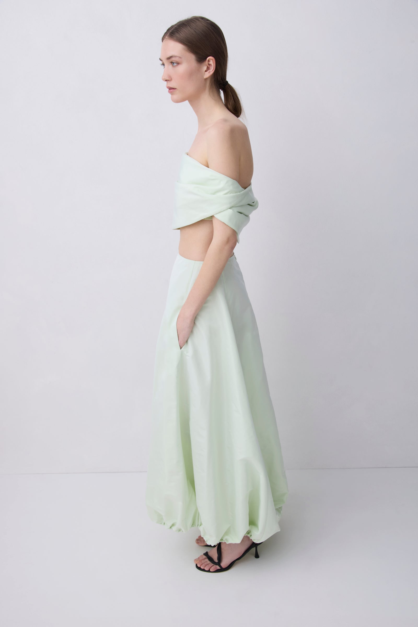 A woman stands in profile wearing a pale mint green two-piece outfit with an off-the-shoulder wrap-style crop top and a matching long skirt, paired with black strappy heels, against a plain light gray studio background.