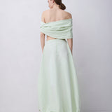 A woman stands with her back to the camera wearing a pale mint green off-the-shoulder maxi dress with a voluminous gathered skirt and black high-heeled sandals, set against a plain light gray studio background.