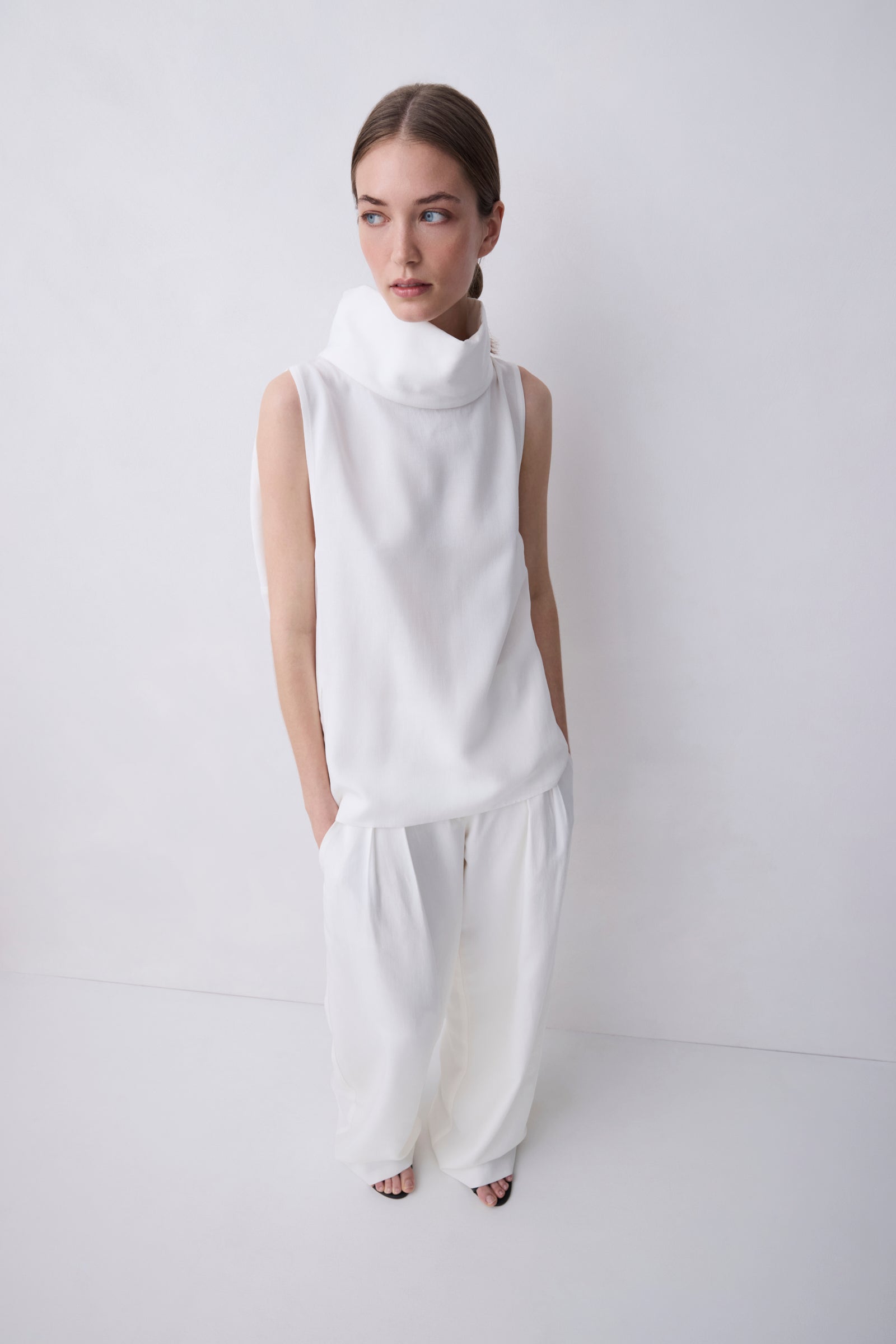 A model wearing a white sleeveless mock-neck top and matching wide-leg trousers, standing against a plain white studio background.