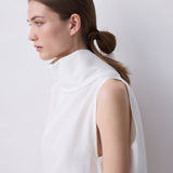 A woman in profile wearing a white sleeveless mock-neck top, photographed against a plain light gray studio background.