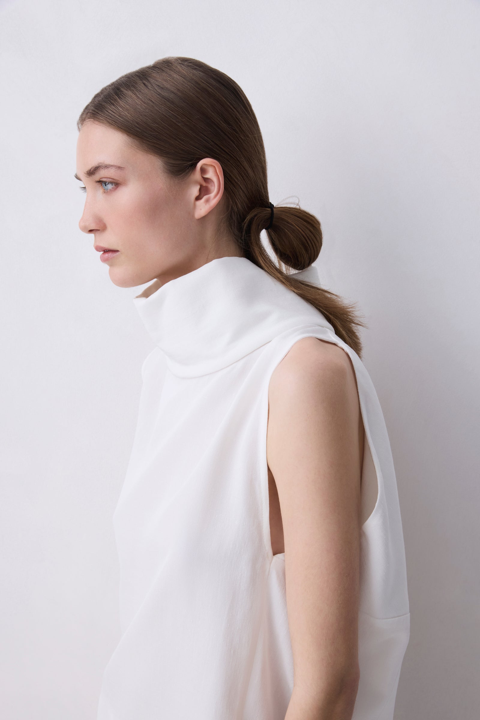 A woman in profile wearing a white sleeveless mock-neck top, photographed against a plain light gray studio background.