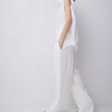 A model wearing a white sleeveless high-neck jumpsuit with wide-leg trousers, standing in profile against a plain white studio background.