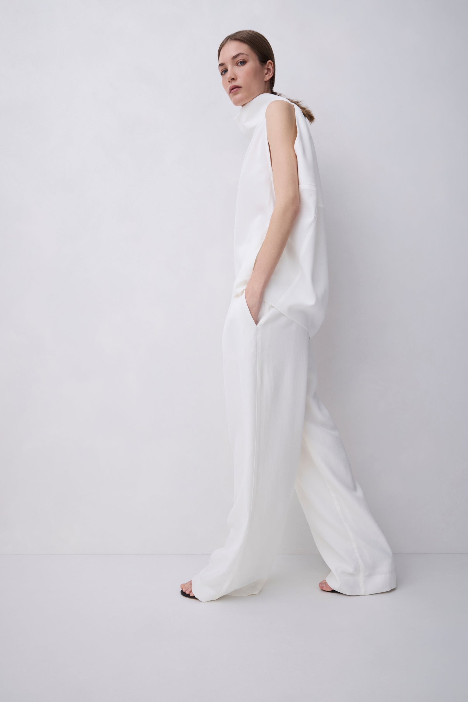 A model wearing a white sleeveless high-neck jumpsuit with wide-leg trousers, standing in profile against a plain white studio background.