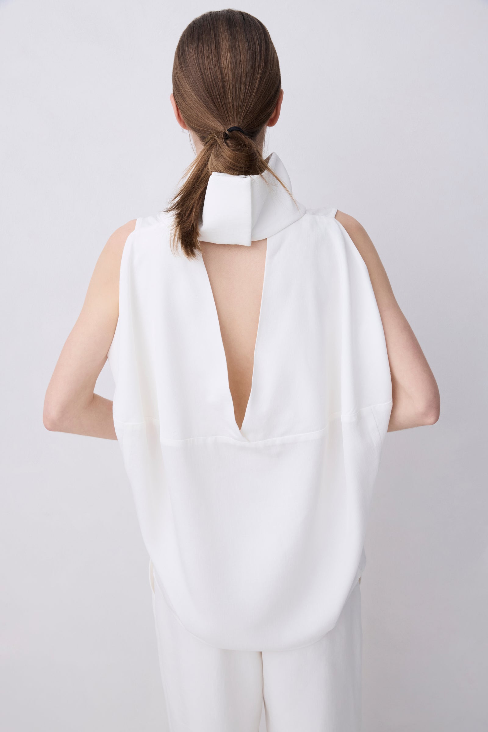 A model shown from the back wearing a white sleeveless top with a high structured collar and a deep V-back cutout, paired with matching white trousers, against a light gray studio background.