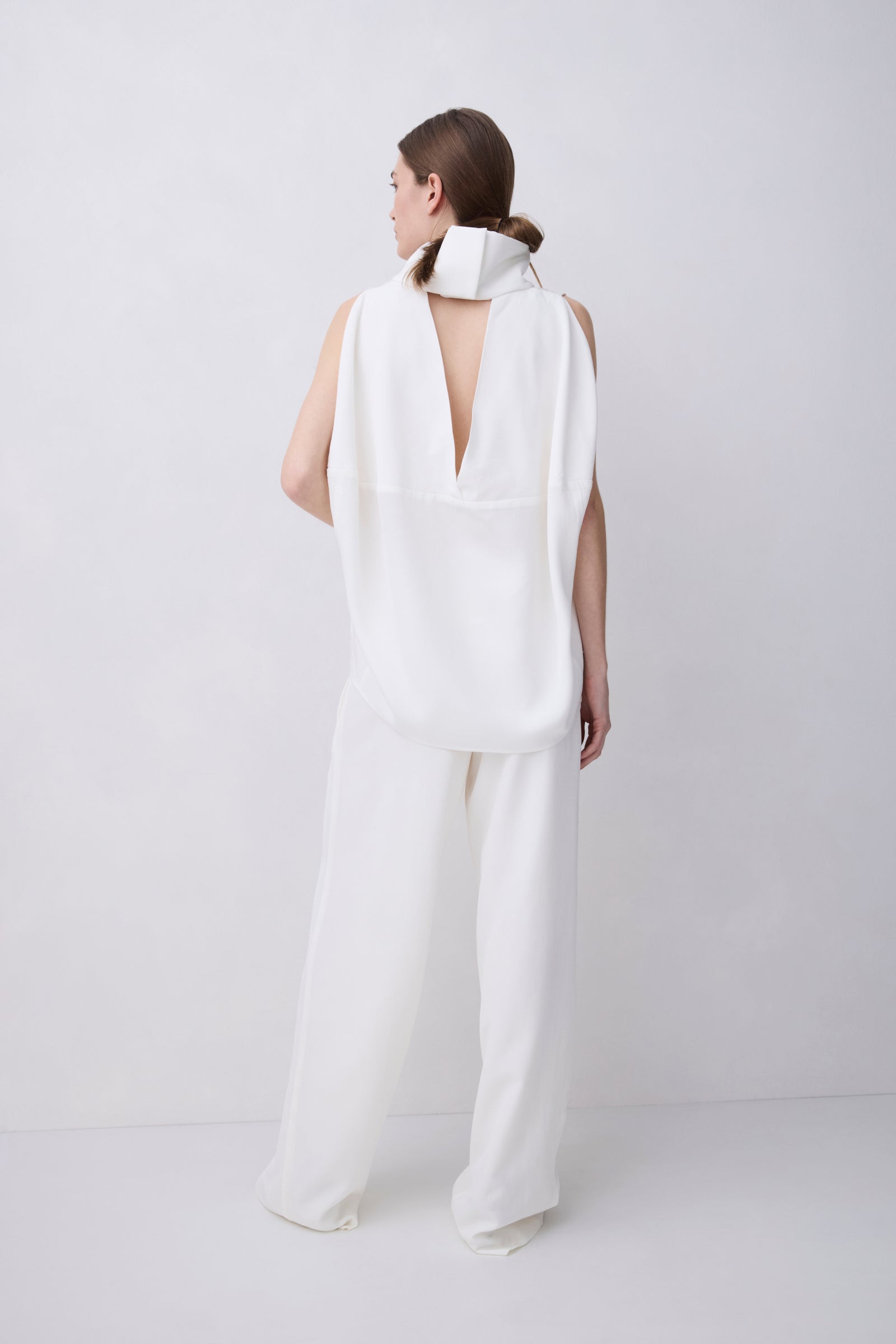 A model shown from the back wearing a white sleeveless jumpsuit with a high structured collar and a deep open back, paired with matching wide-leg trousers, set against a plain light gray studio background.