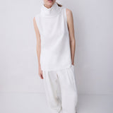 A model wearing a white sleeveless mock-neck top and matching white wide-leg trousers, standing against a plain light gray studio background.