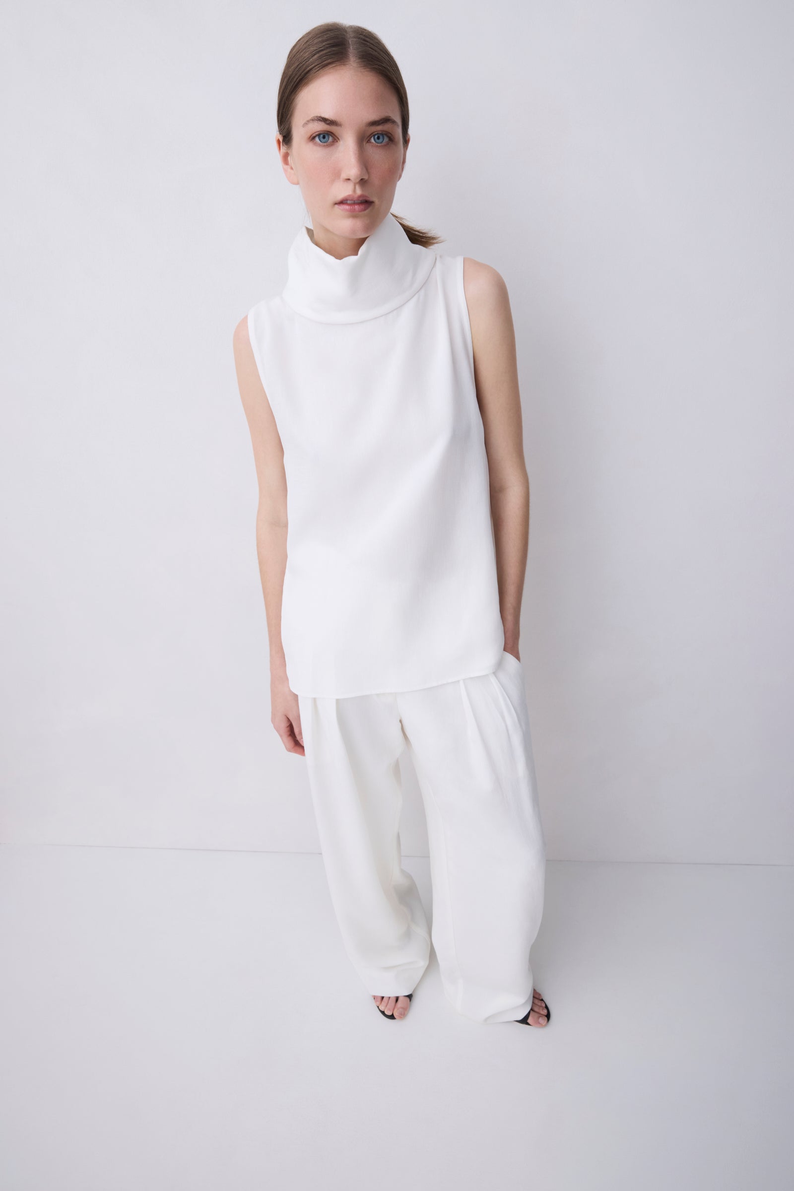 A model wearing a white sleeveless mock-neck top and matching white wide-leg trousers, standing against a plain light gray studio background.