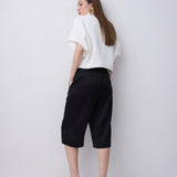 A woman stands with her back to the camera wearing a white short-sleeve blouse, black cropped pants, and black heeled sandals against a plain light gray studio background.