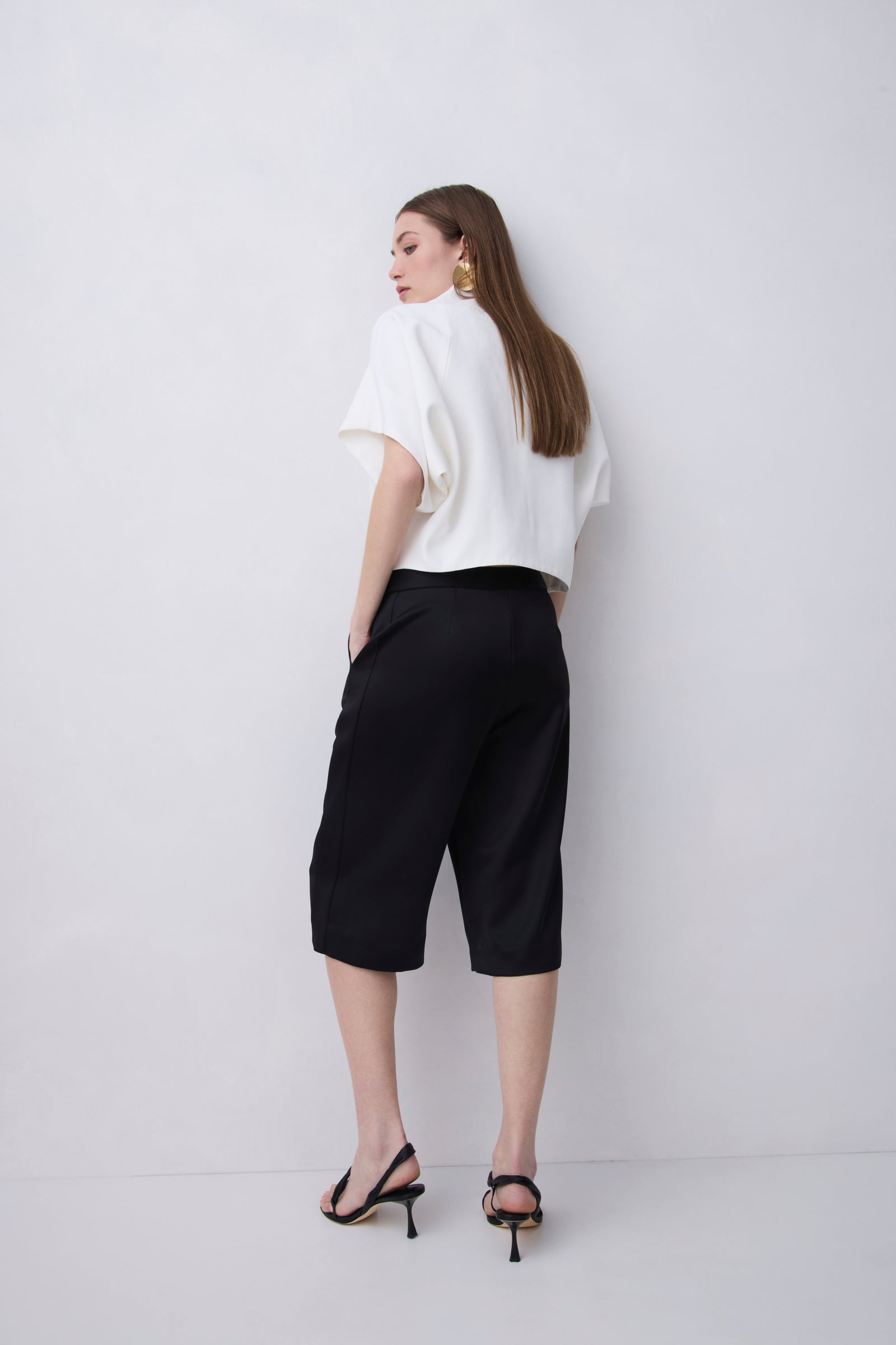 A woman stands with her back to the camera wearing a white short-sleeve blouse, black cropped pants, and black heeled sandals against a plain light gray studio background.