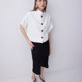 A woman modeling a white short-sleeve blouse with a high mock neck and black buttons, paired with black midi skirt and black strappy heels, against a plain white studio background.