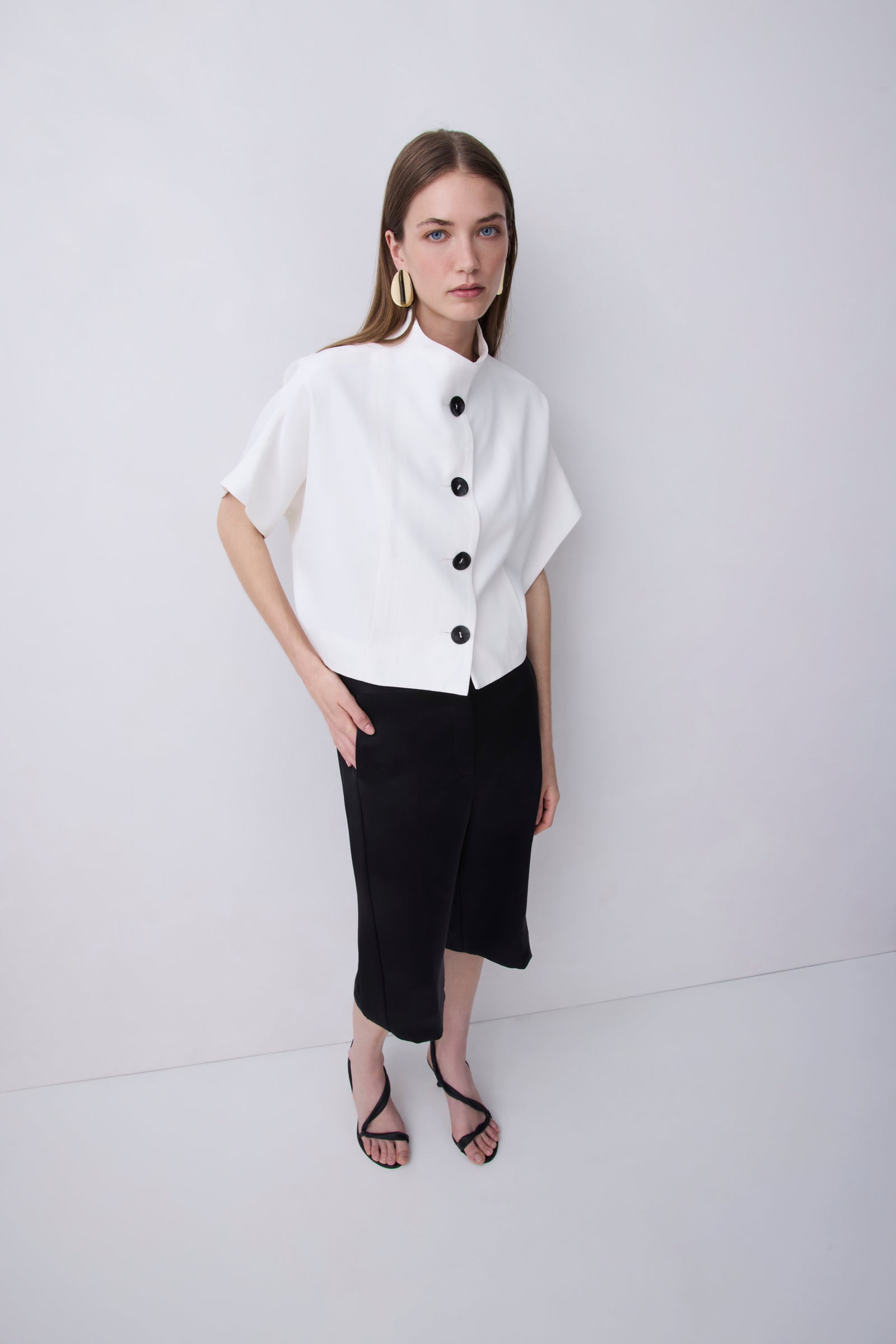 A woman modeling a white short-sleeve blouse with a high mock neck and black buttons, paired with black midi skirt and black strappy heels, against a plain white studio background.