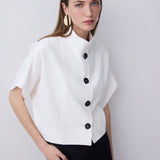 A woman modeling a white short-sleeve cropped jacket with a high stand collar and black buttons, paired with black trousers, against a light gray studio background.