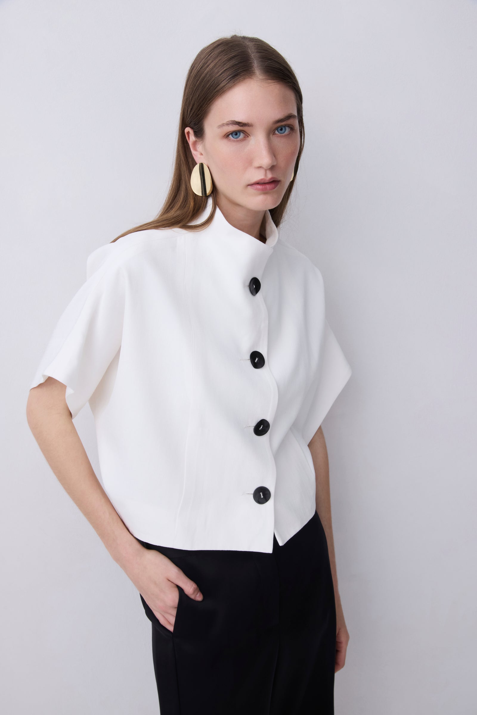 A woman modeling a white short-sleeve cropped jacket with a high stand collar and black buttons, paired with black trousers, against a light gray studio background.