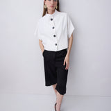 A woman modeling a white short-sleeve blouse with black buttons and a high mock neck, paired with black midi skirt pants and black strappy heels, against a light gray studio background.