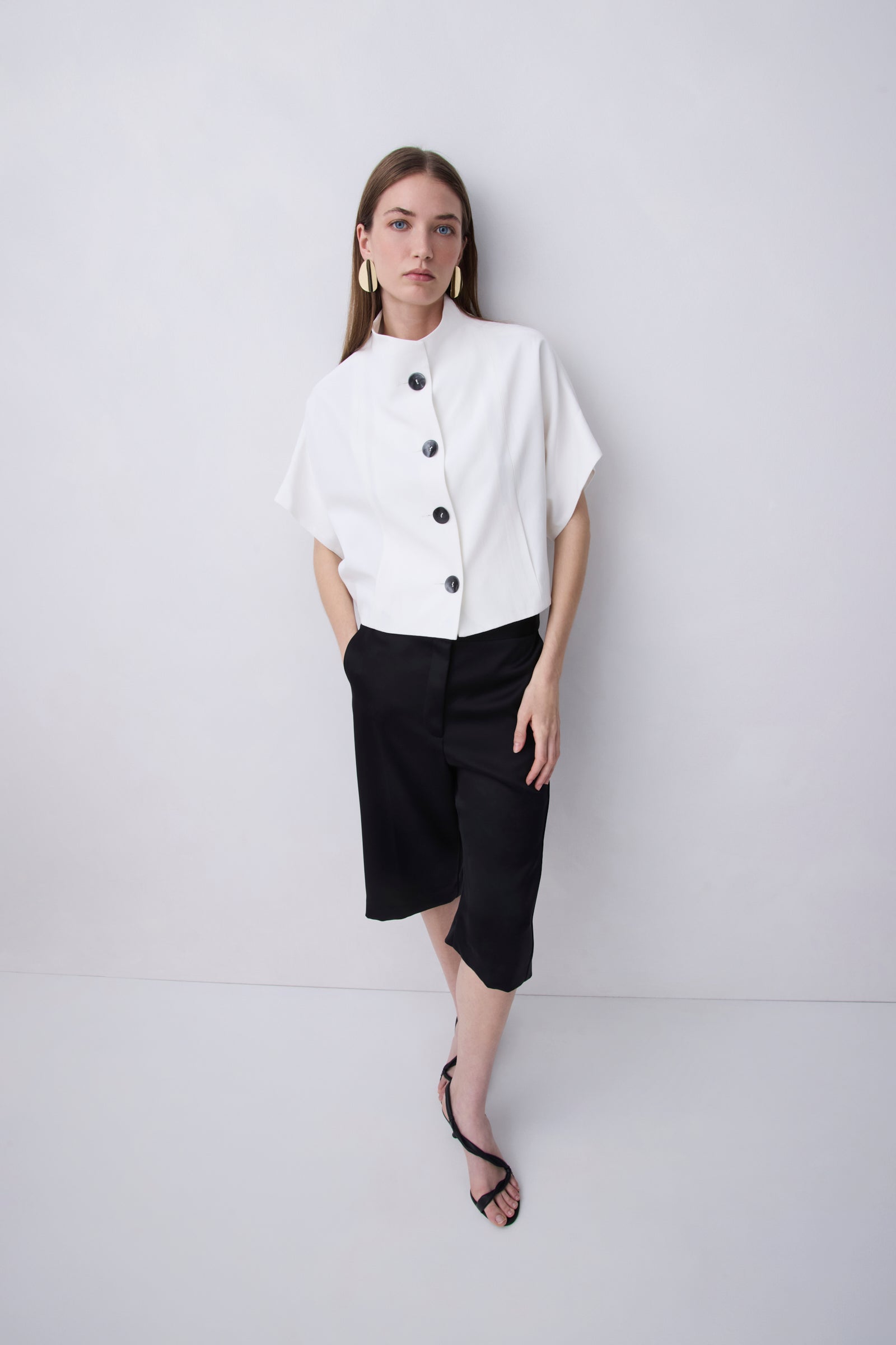 A woman modeling a white short-sleeve blouse with black buttons and a high mock neck, paired with black midi skirt pants and black strappy heels, against a light gray studio background.