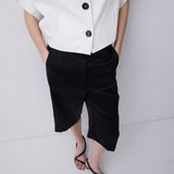 A model wearing a white short-sleeve button-up shirt and black cropped shorts, paired with black strappy sandals, against a light gray studio background.