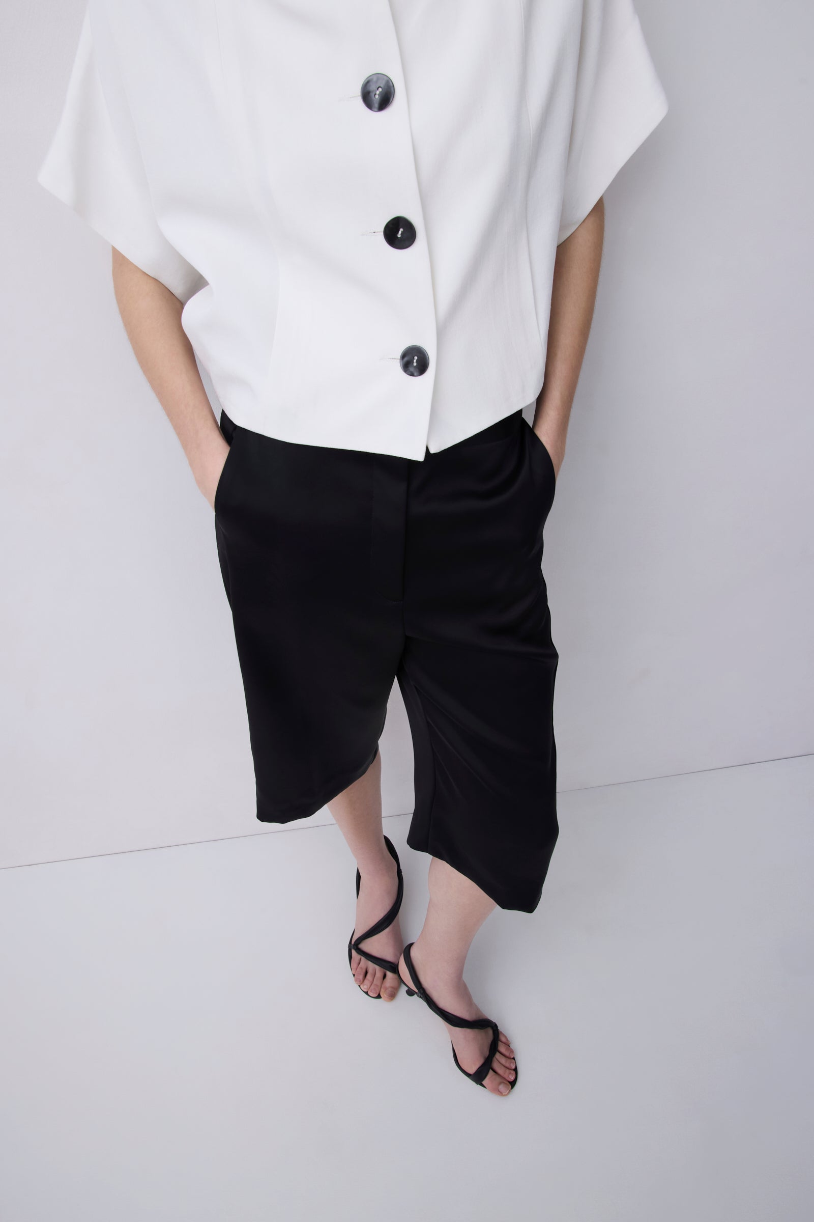 A model wearing a white short-sleeve button-up shirt and black cropped shorts, paired with black strappy sandals, against a light gray studio background.
