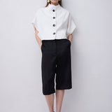 A woman stands against a light gray studio wall wearing a white short-sleeve blouse with a high mock neck and black buttons, paired with black cropped pants and black strappy sandals.