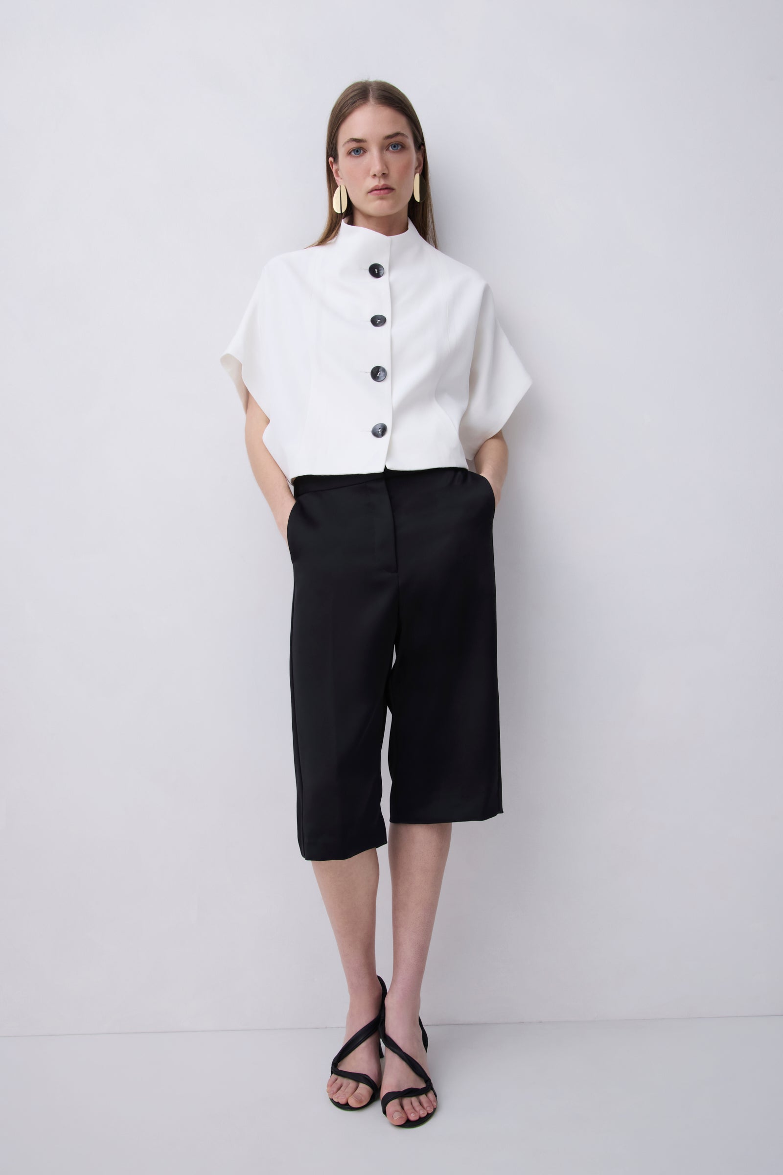 A woman stands against a light gray studio wall wearing a white short-sleeve blouse with a high mock neck and black buttons, paired with black cropped pants and black strappy sandals.