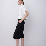 A woman modeling a white short-sleeve button-up shirt and black cropped midi skirt, paired with black strappy high-heeled sandals, against a light gray studio background.