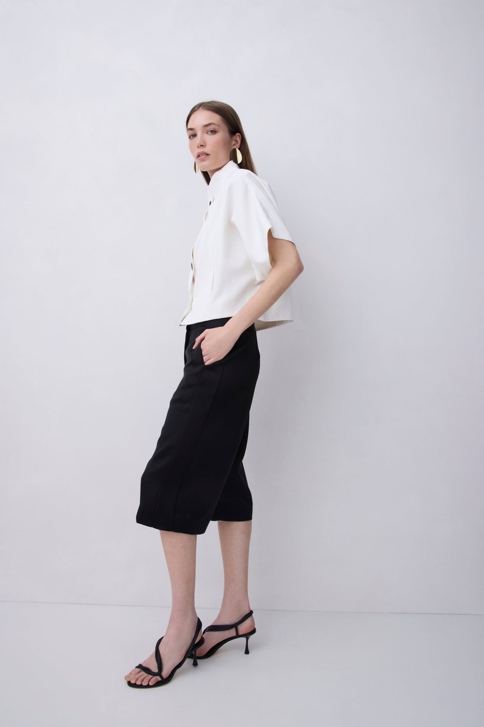 A woman modeling a white short-sleeve button-up shirt and black cropped midi skirt, paired with black strappy high-heeled sandals, against a light gray studio background.