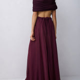 A woman modeling a deep burgundy two-piece outfit consisting of an off-the-shoulder wrap crop top and a matching high-waisted, flowing maxi skirt, set against a plain light gray studio background.