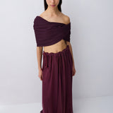A model wearing a deep plum purple two-piece outfit consisting of an off-the-shoulder wrap top and a matching long pleated skirt, standing against a light gray studio background.