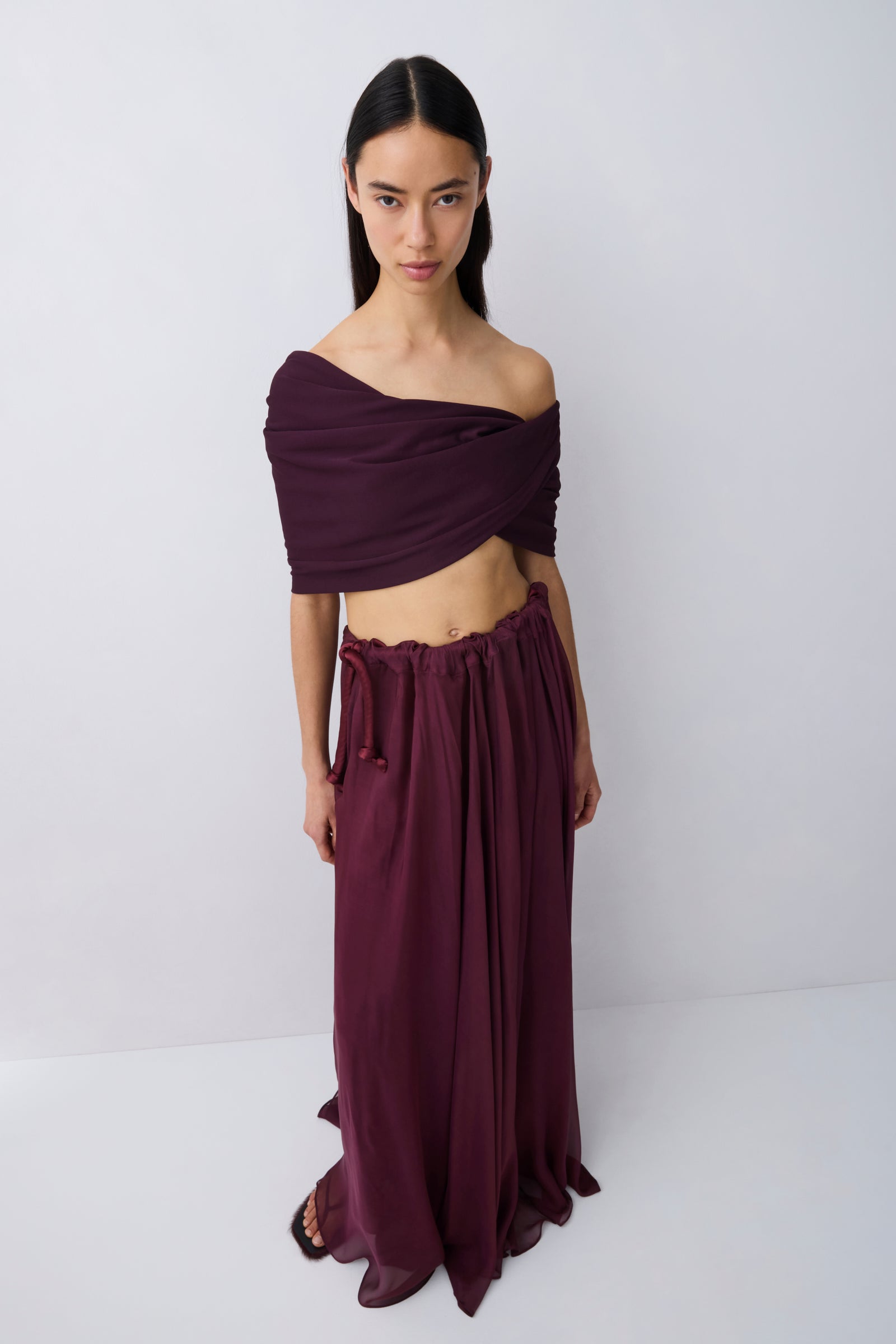 A model wearing a deep plum purple two-piece outfit consisting of an off-the-shoulder wrap top and a matching long pleated skirt, standing against a light gray studio background.