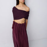 A woman modeling a deep burgundy two-piece outfit consisting of an off-the-shoulder wrap crop top and a matching high-waisted, flowing maxi skirt, set against a plain light gray studio background.
