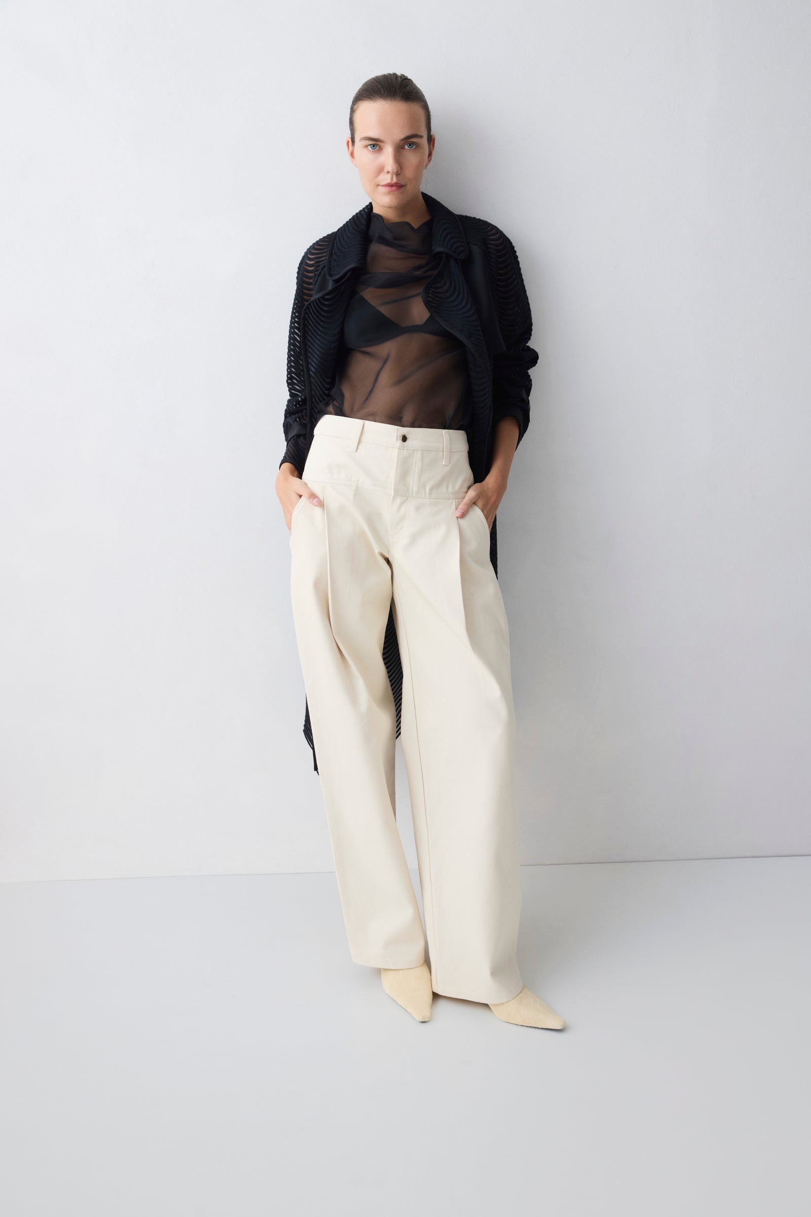Person wearing a black sheer top, black cardigan, and white pants against a white background